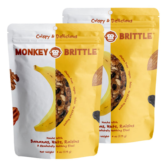 Two yellow and white resealable bags of monkey brittle on a white background. The package features a banana in the center and nuts and raisins on the edge. Crispy and delicious is written at the top of the package